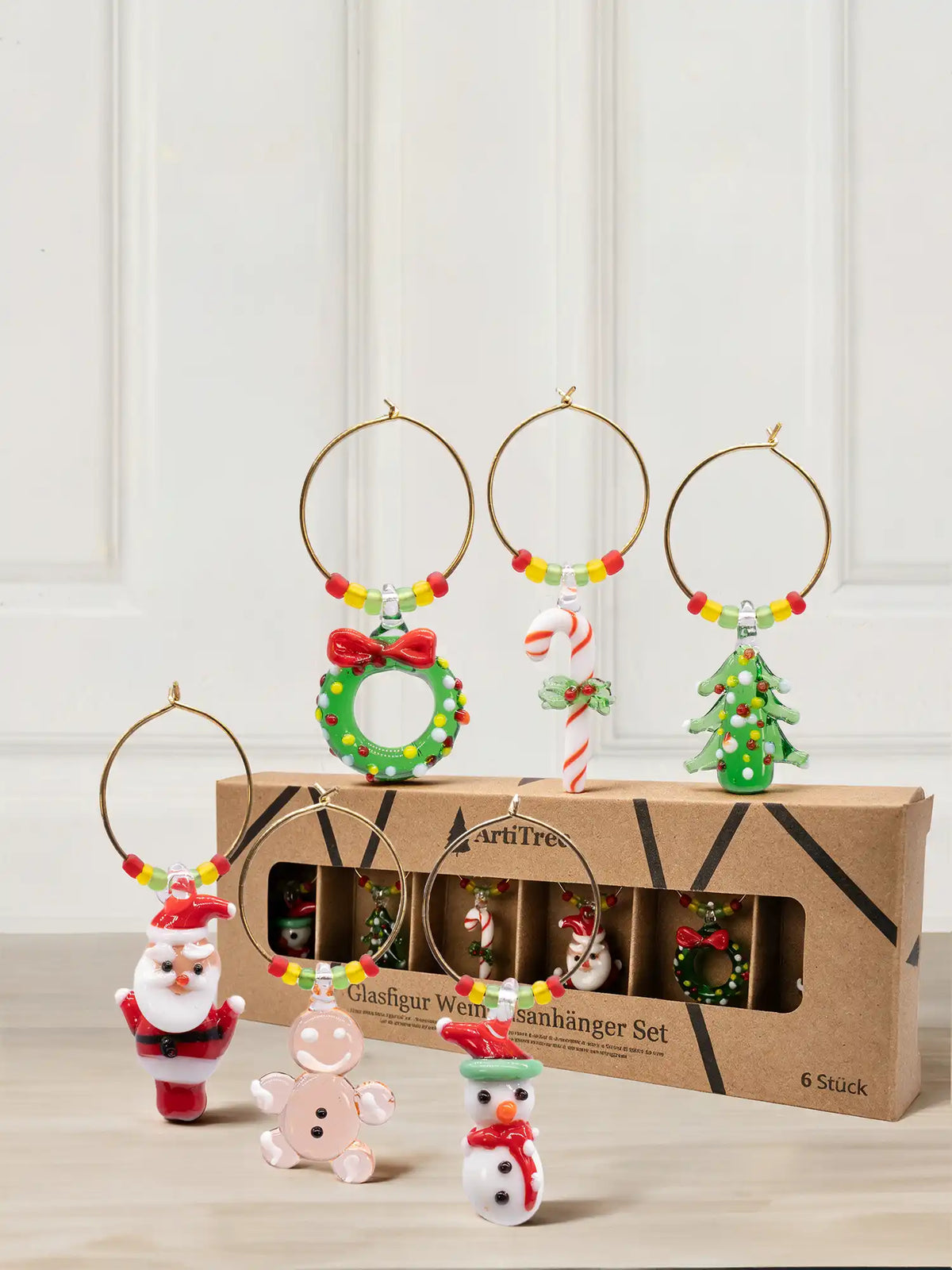 Set of six wine glass charms with gold-tone rings and colorful beads: Santa, gingerbread man, snowman, wreath, candy cane, and glass Christmas tree in front of a cardboard box with a window.