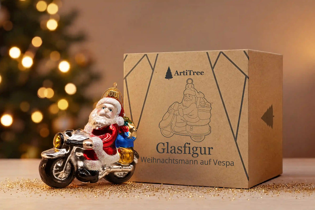 Glazen kerstornament van de kerstman op een scooter met een zak cadeaus, naast de kartonnen ArtiTree-verpakking met het opschrift „Glazen kerstman op Vespa“, met op de achtergrond een verlichte kerstboom