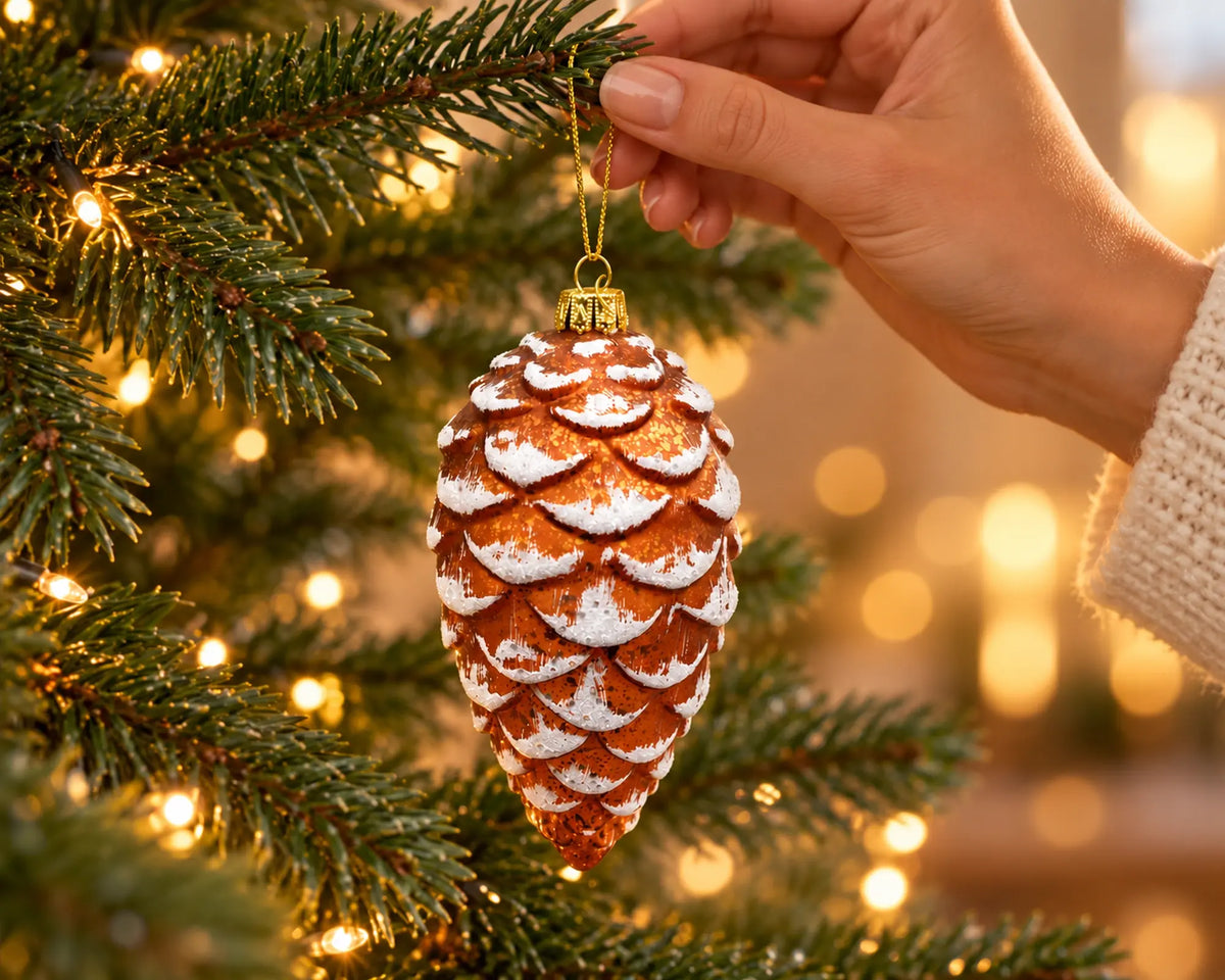 Hand hangt een glazen kerstornament in de vorm van een dennenappel in een verlichte kerstboom, koperbruin met wit besneeuwde schubben en een gouden koord