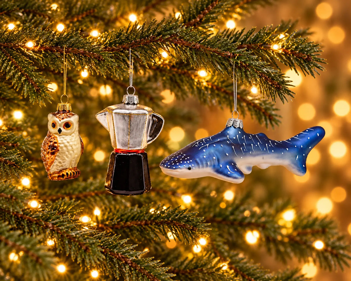 Drie glazen figuren hangen naast elkaar in groottevergelijking aan de verlichte kerstboom: een kleine uil, een middelgrote espressomaker en een lange walvishaai